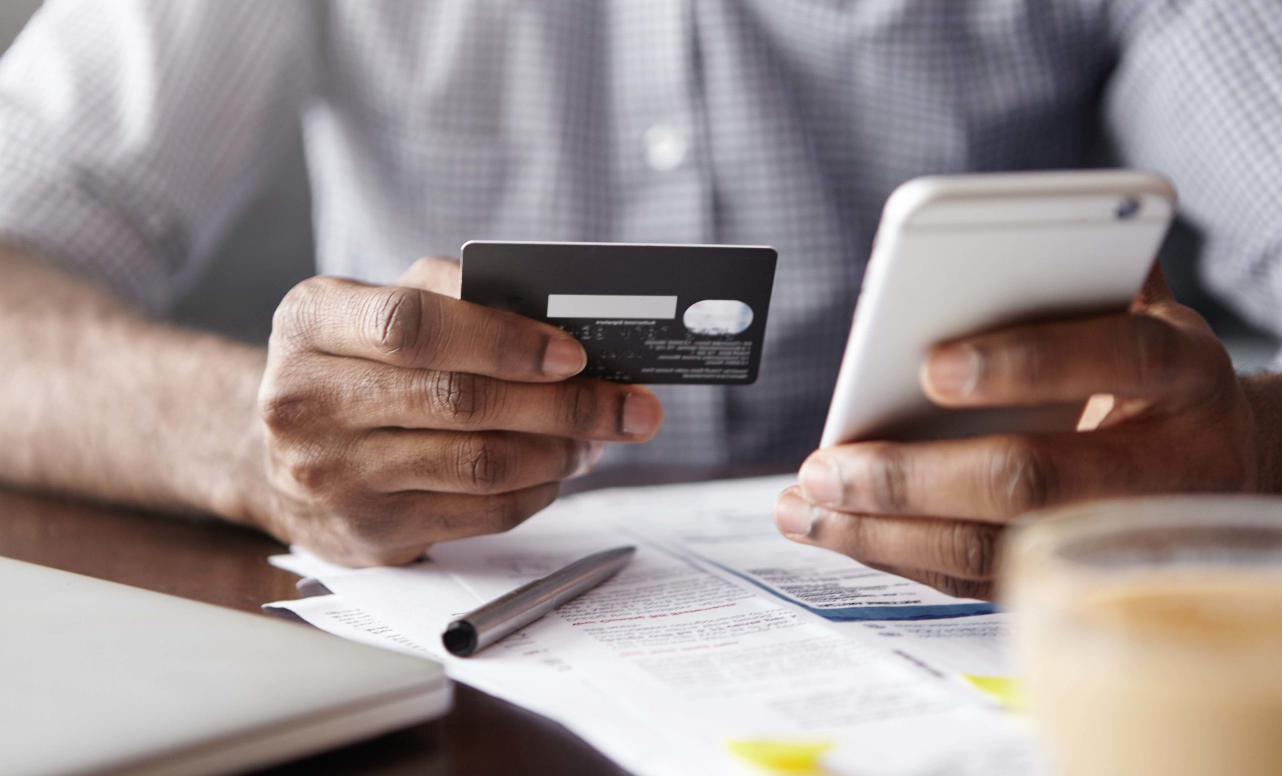 Close-up view of African man's hands holding plastic credit card and smart phone. Dark-skinned businessman using mobile banking application on his gadget while paying bills at cafe online via internet